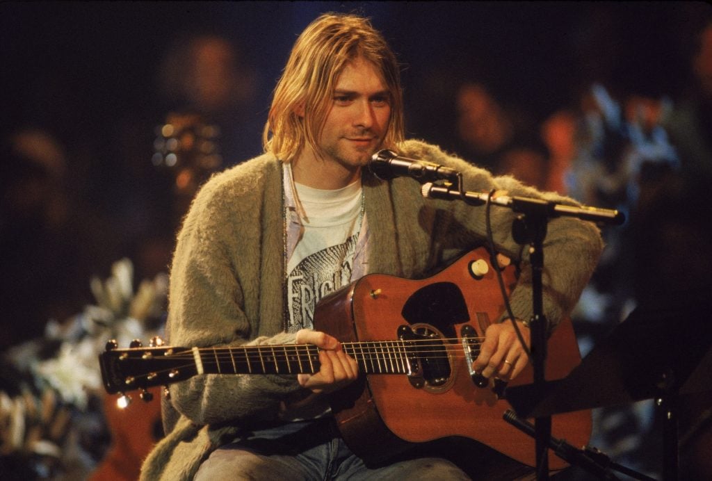 A blonde-haired man, wearing a casual cardigan and graphic t-shirt, plays an acoustic guitar while seated in front of a microphone, performing live on stage