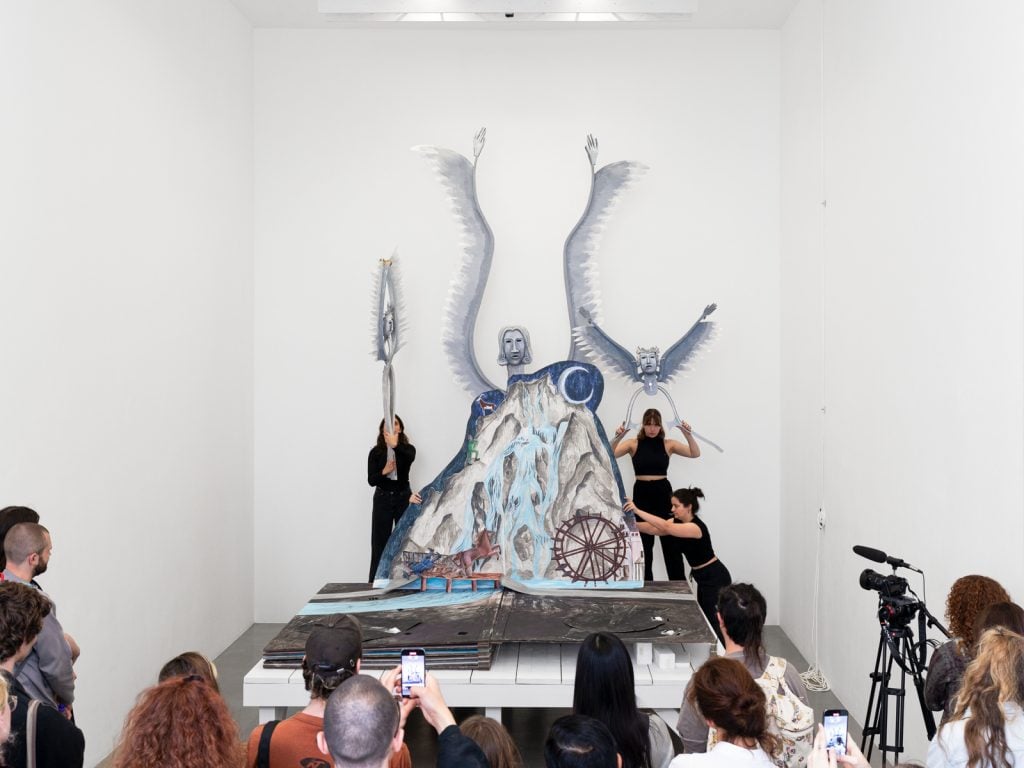 A group of people as audience watching a diorama pop up with performers carrying props, all dressed in black.