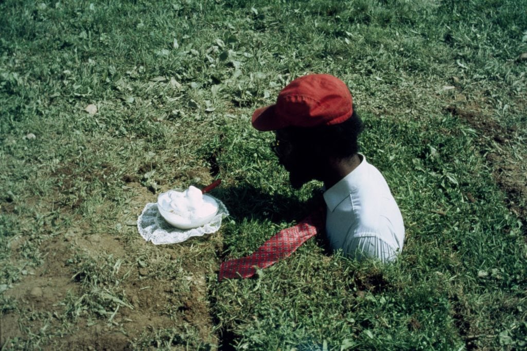 Vintage color photo of a man shoulders and up wearing a red baseball hat and white tee emerging from a patch of grass in profile facing a plate with a whipped cream dessert, featured at the salon by nada.