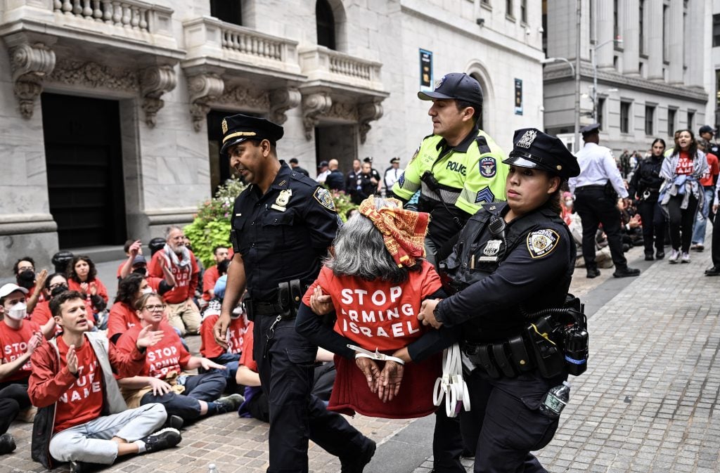 An elderly woman being carried away by three police officers while protesters in red t-shirts sit on the ground in the background