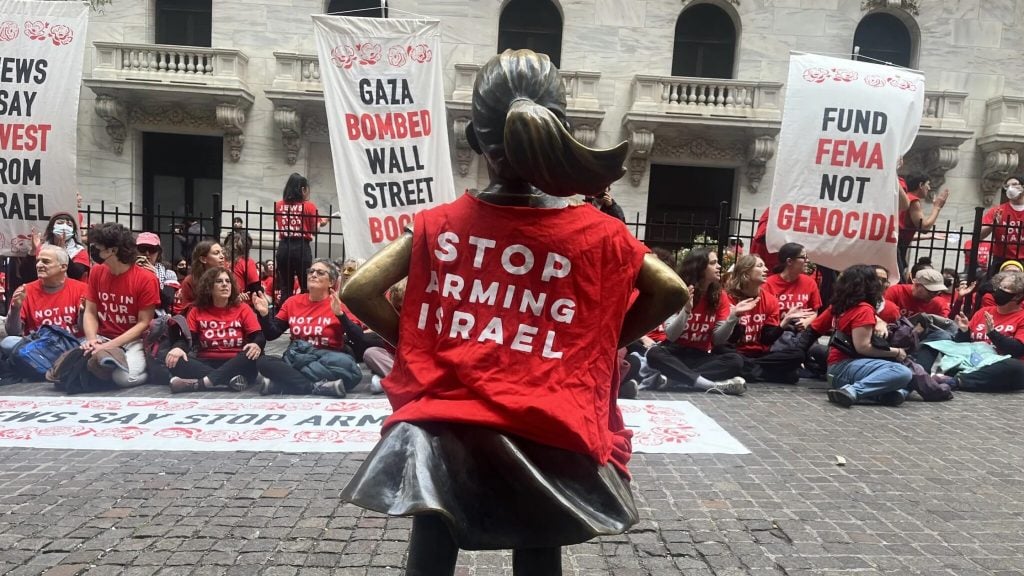 A photo of a sculpture of a little girl facing the Stock Exchange wearing a t shirt reading "stop arming Israel"