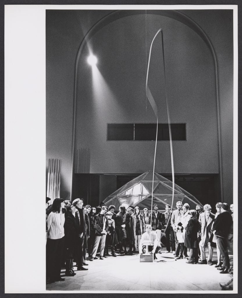 A crowd gathered around a man crouching on the ground an activating a device that is shooting a ribbon into the air