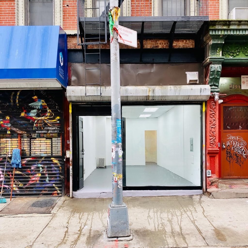 The Satellite Art Show gallery on Broome Street on New York's Lower East Side. An empty, white walled gallery in a storefront is seen from the sidewalk behind a lamppost with a parking regulation sign. 