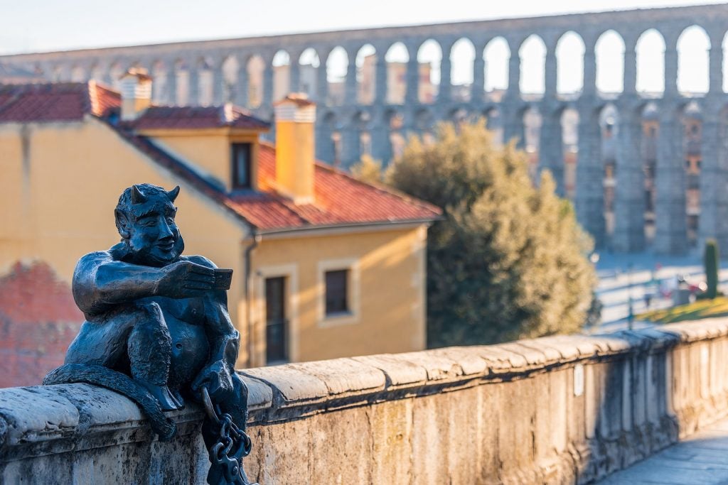 A bronze statue of a devil taking a selfie perched on a stone ledge with the Segovia Aqueduct in the background