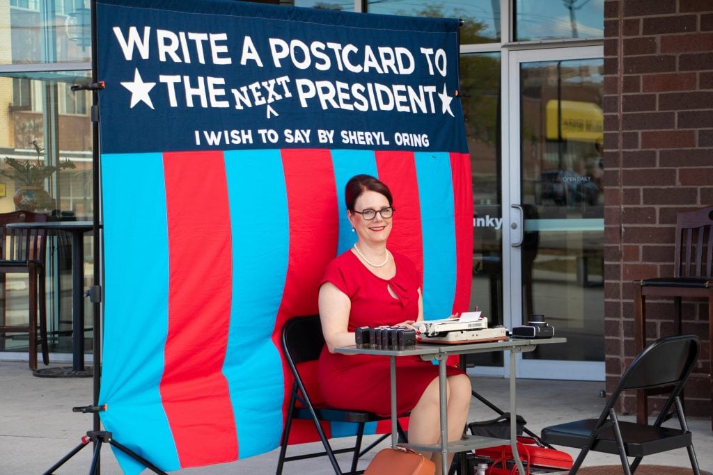 Sheryl Oring performing I Wish to Say at Gem City Market in Dayton, Ohio. She is wearing a vintage red dress and pearls, like a 1950s secretary, and is seated in front of a red and blue striped banner inviting the public to write a letter to the next president.