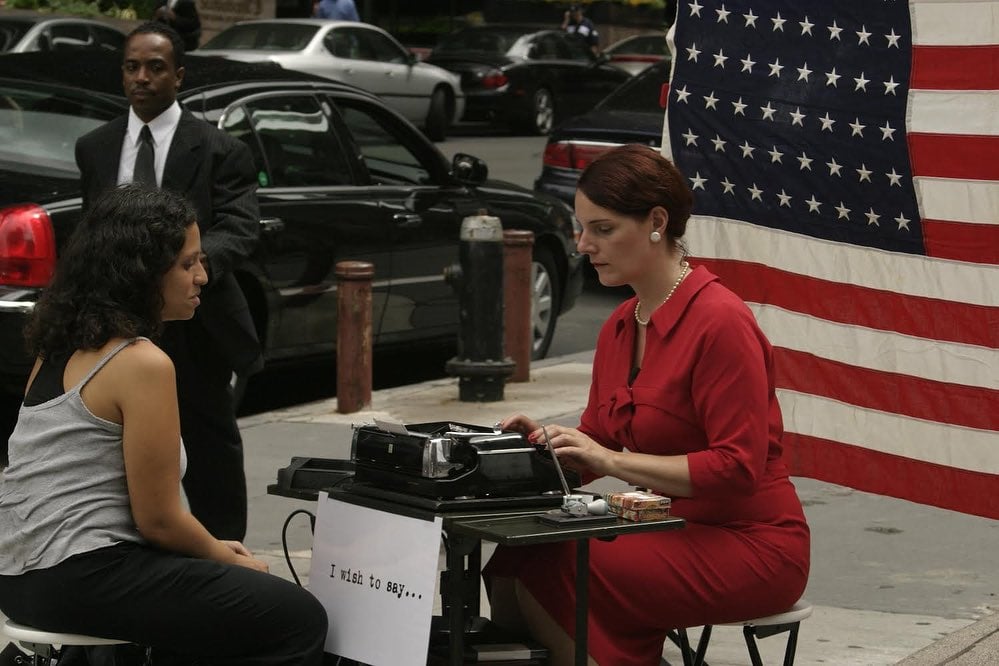 Sheryl Oring performing <em>I Wish to Say</em> during the 2004 Republican National Convention in New York. The artist, dressed as a 1950s secretary, sits at a typewriter in front of an American flag. 