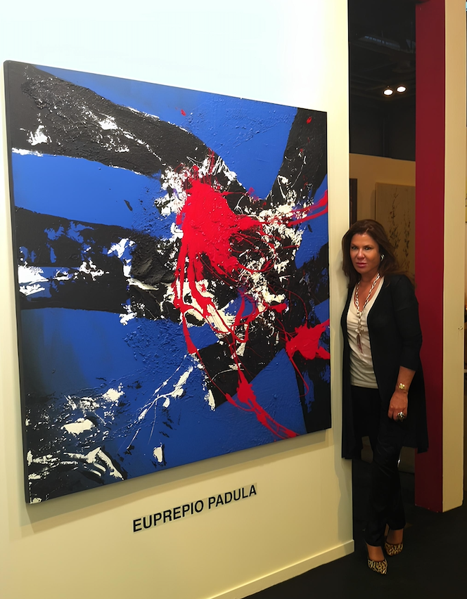 Gallerist Soraya Cartategui standing beside an abstract painting in blue, red, white, and black.