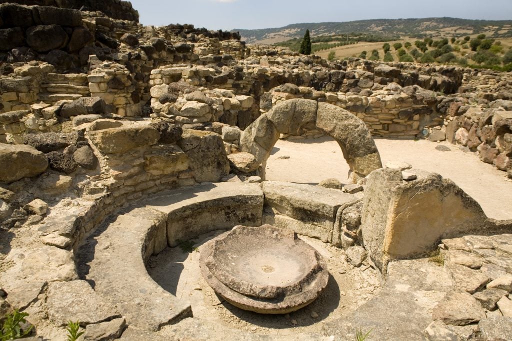 a collection of old rocks arranged around a well surrounded by pillars and low walls under a blue sky