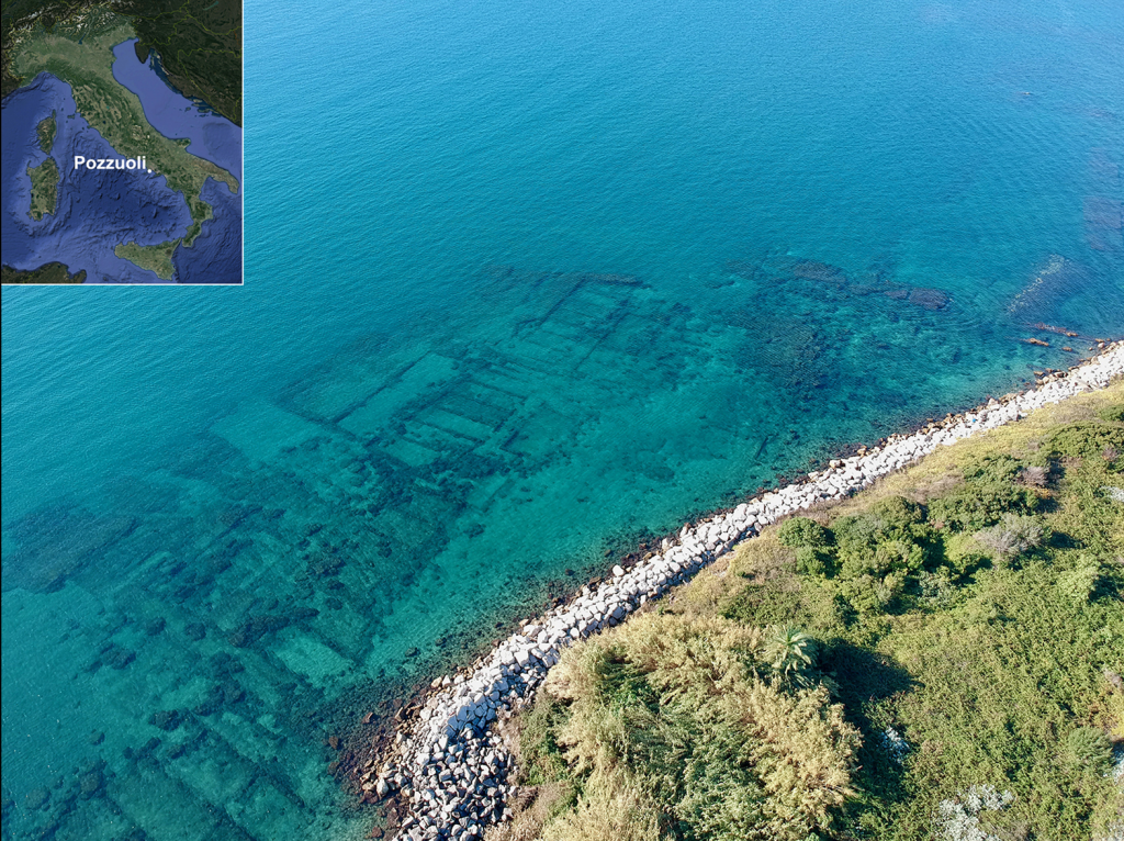 a group of old buildings submerged beneath clear blue water close to land with grass and rocks