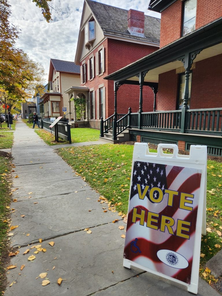 A "vote here" sign outside the Susan B. Anthony Museum and House, a two-story brick building with a porch on a residential block in Rochester, New York.