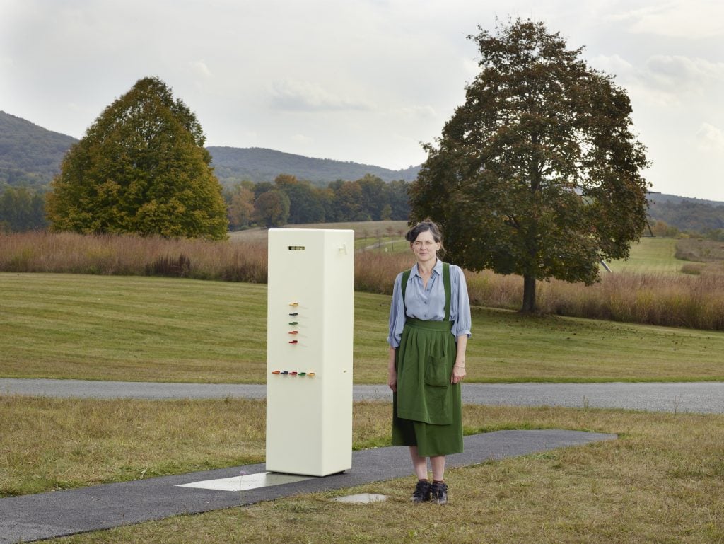 Artist Taryn Simon standing next to her Kleroterion-inspired sculpture machine, which is epoxy white with colorful resin bars sticking out of the front and set agains a rural Northeastern landscape.