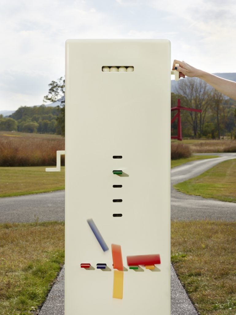 A slow motion photo of the Kleroterion machine by Taryn Simon with a hand reaching to pull a lever and the four of five colorful resin bars falling out of the front of the machine.