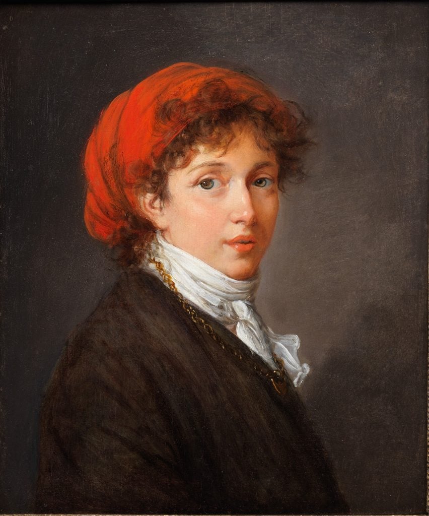 A self-portrait of a woman in dark jacket and a red head wrap against a dark background