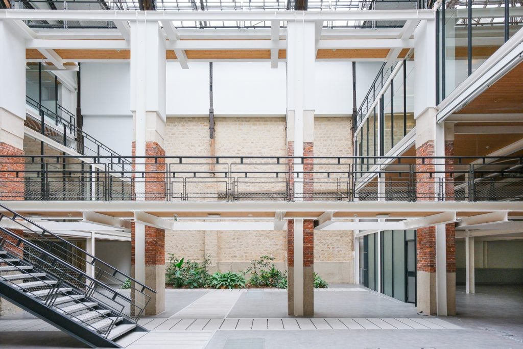 Inside the industrial space in paris where the salon by nada will be held, with an atrium glass ceiling, four brick and plaster columns, and a steel staircase leading up to a second floor on the left.