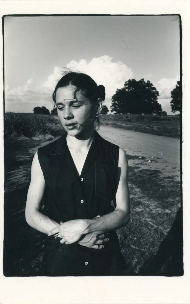 Gerard E. Murrell, On the Cane Break: Portrait of Tina Girouard, Cecilia, Louisiana (1976). A black and white photo of a young woman with dark hair pulled back and a dark sleeveless v-neck button-down shirt stands, photographed from the waist up with her arms crossed below her breasts, in a field in Louisiana.