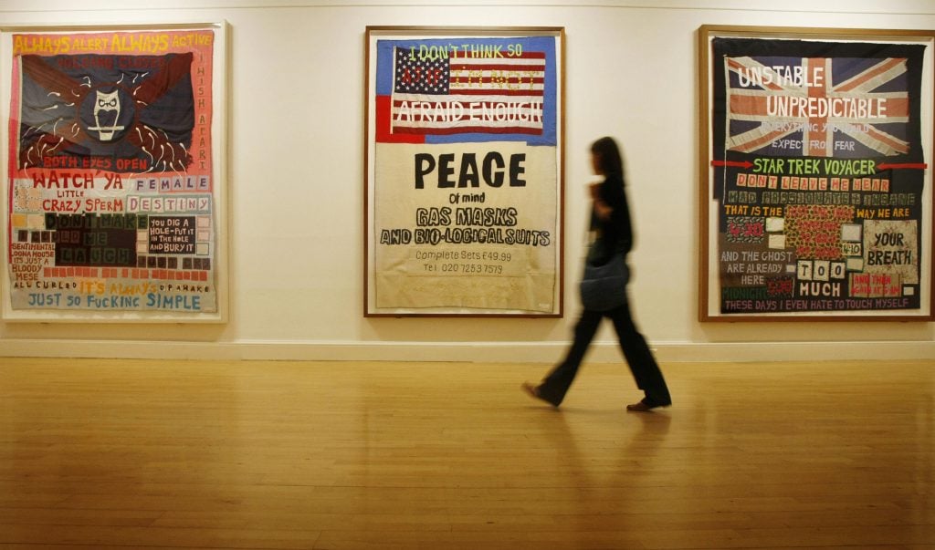 A woman walking past three framed blankets, sewn with phrases and patterns, hanging in a gallery.