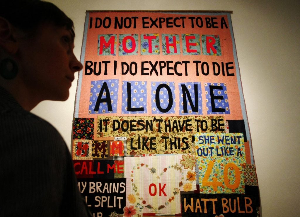 A person silhouetted against a large framed blanket sewn with the words 'I do not expect to be a mother but I do expect to die alone.'