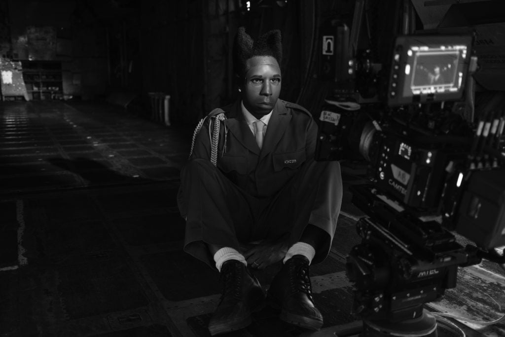 Black and white photo of a man in a full face mask and suit sitting on the ground while looking into a camera