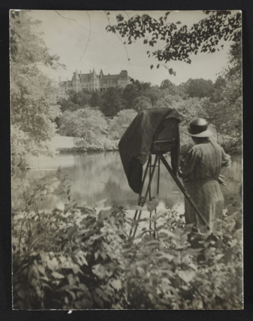 a black and white photograph of a man taking a photograph of a large mansion atop a hill surrounded by trees