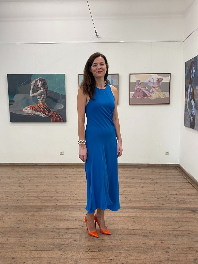 Full-length portrait of Virginie Tison, founder of Art Professional School, wearing a long blue dress and orange heels standing in a gallery space with several paintings installed along the walls.