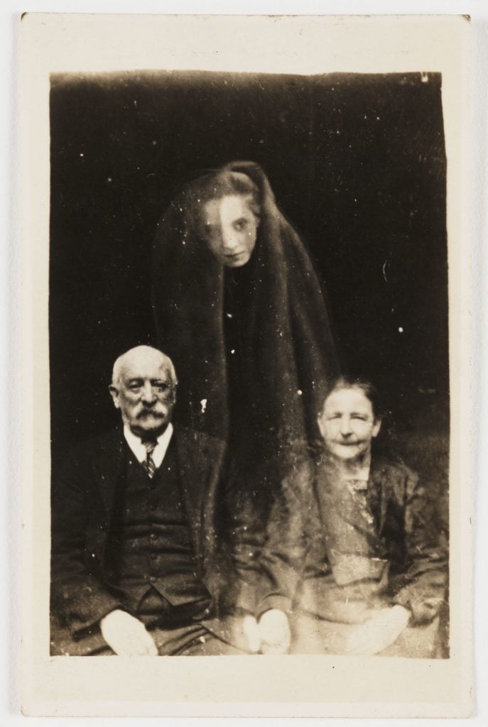 A young woman's face appears as if floating above a seated elderly couple, draped in a cloak.