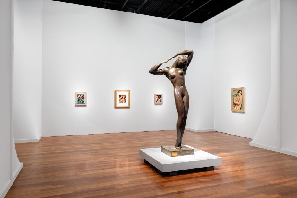 Installation view showing a sculpture of a nude woman with her arms touching behind her head and paintings by Tamara de Lempicka