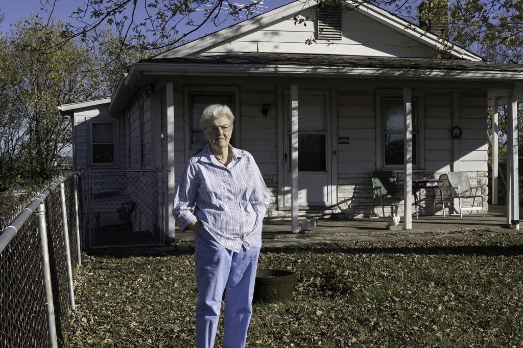 An elderly woman stands in front of a home with a smile and her hands in her pockets.