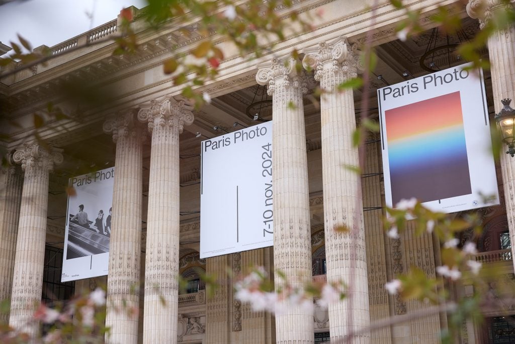 Large banners for "Paris Photo" hang between grand columns of a neoclassical building, promoting the event from November 7-10, 2024, with foliage framing the scene.