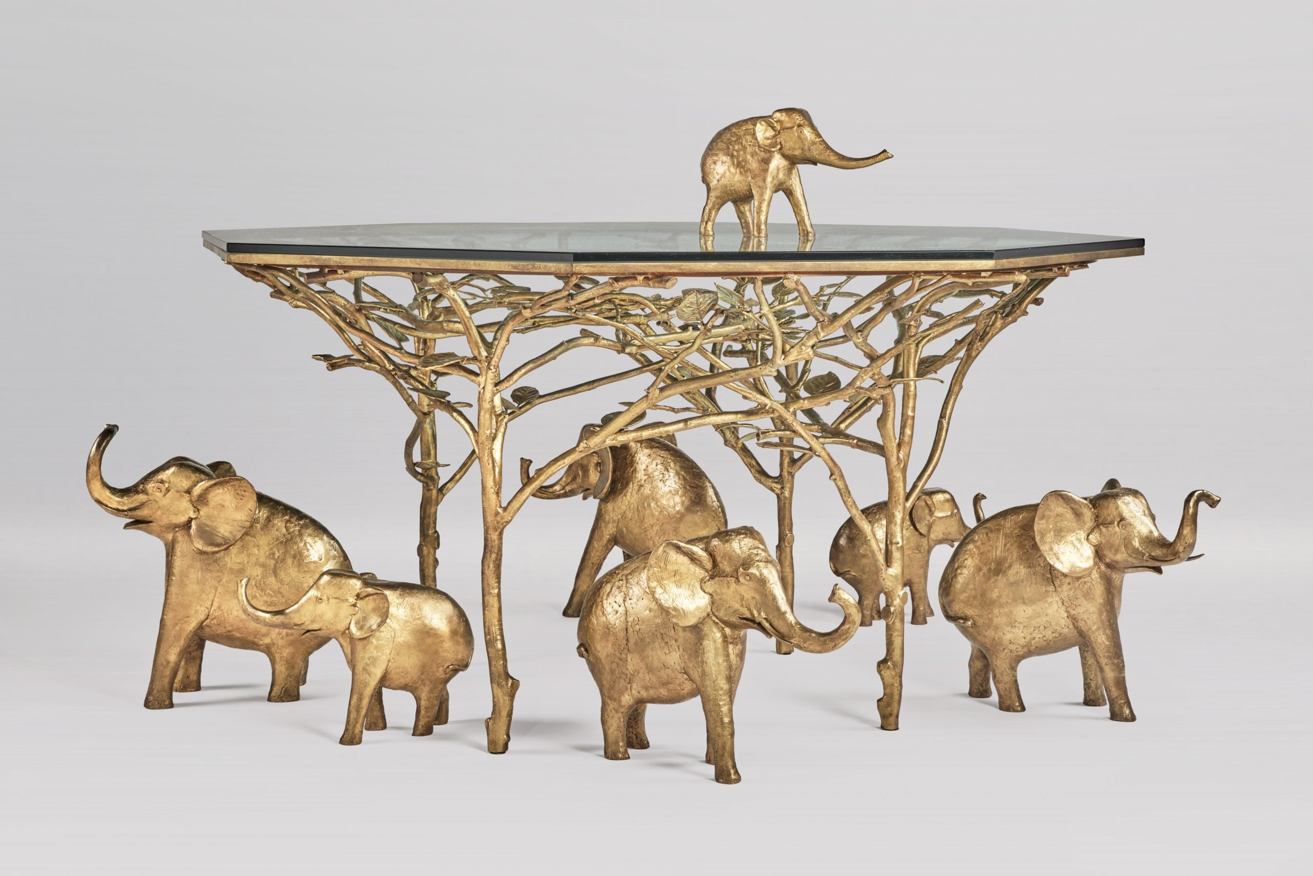 A decorative table with a glass top supported by a sculptural base featuring golden elephant figures intertwined with tree-like branches.