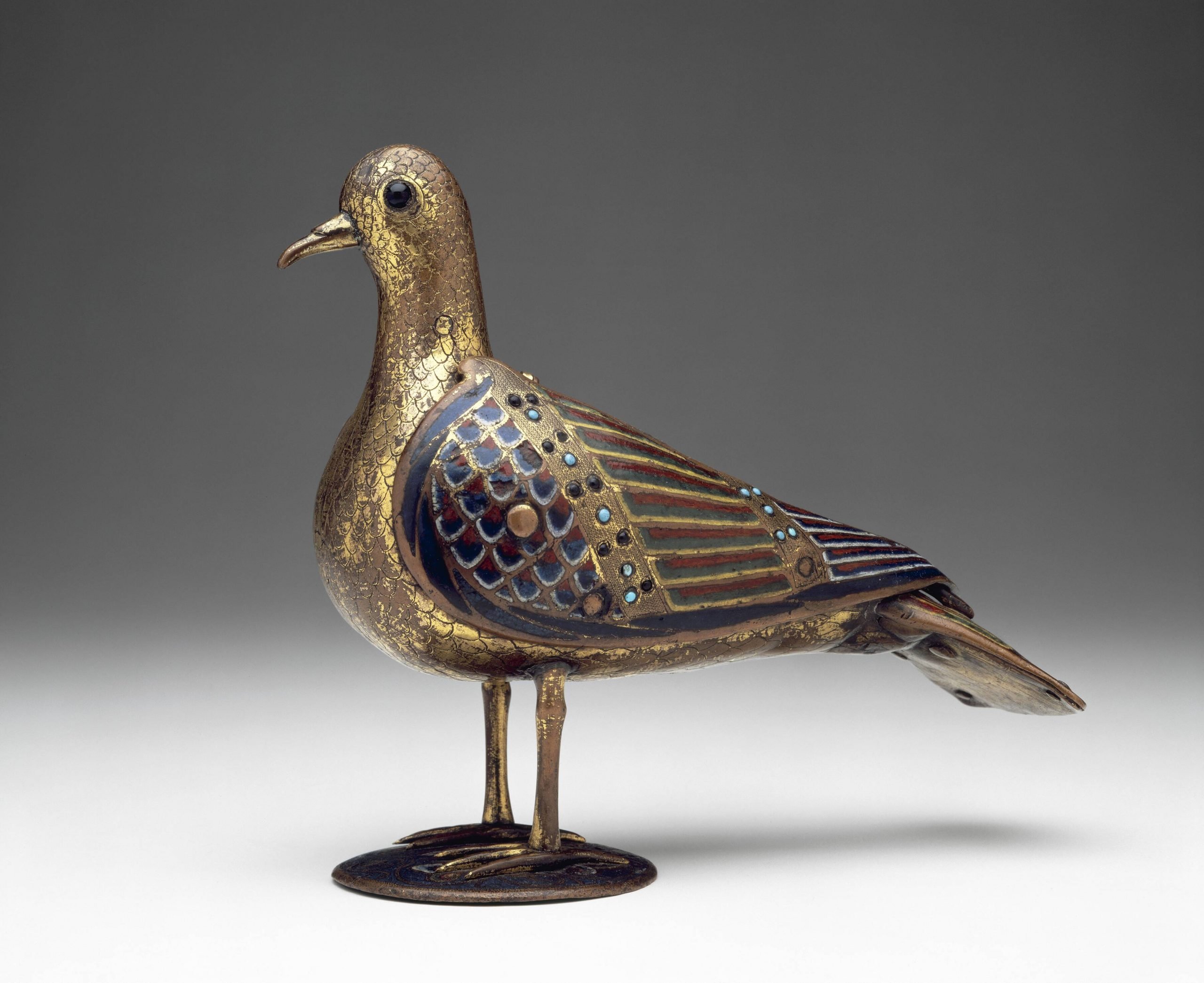 a statuette of a dove with a golden head and brass wings and tail feathers on a small pedastal against a dark background inside an exhibition space