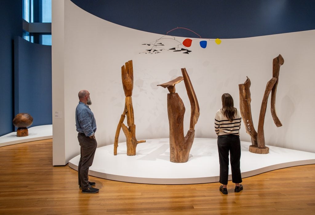 a group of people standing in front of a collection of tall wooden statues on a white stage inside a brightly lit exhibition space