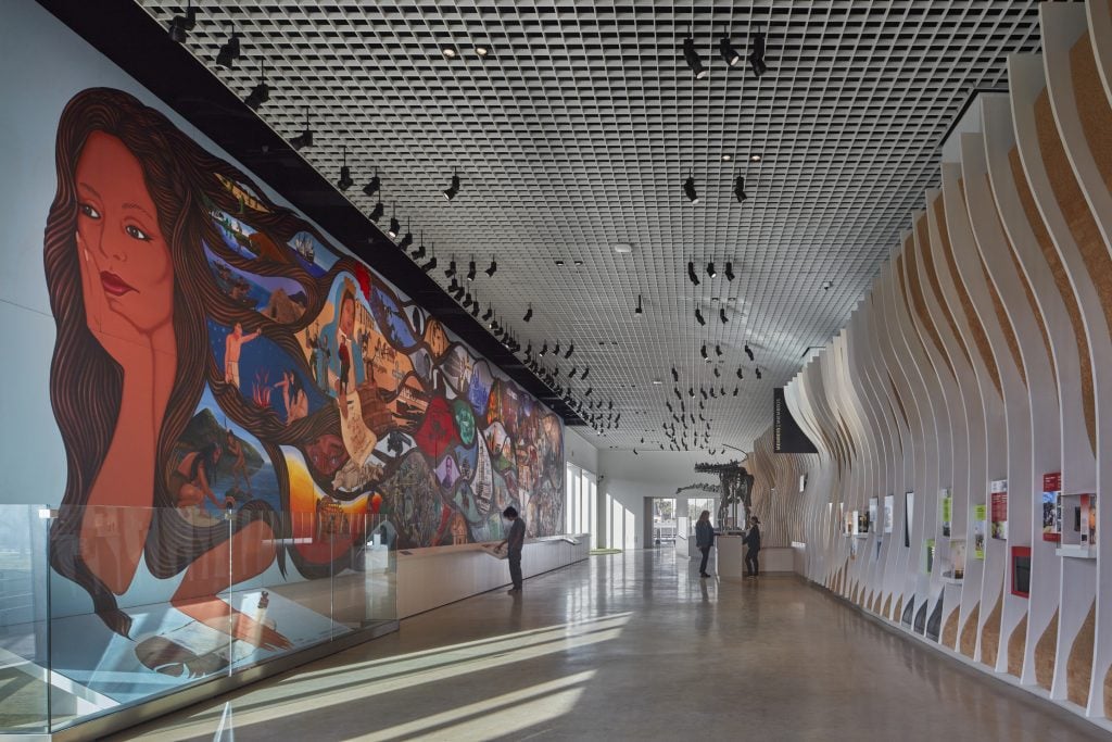 A long gallery at the new wing of the L.A. Natural History Museum, with a massive mural covering one wall and a dinosaur fossil towards the end of the hall