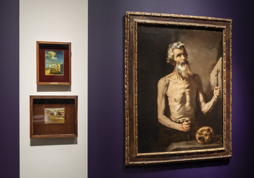 painting of an old man with a bare chest and a long beard spreading his arms inside a fancy frame on a dark purple blue wall next to two smaller images inside an exhibition space