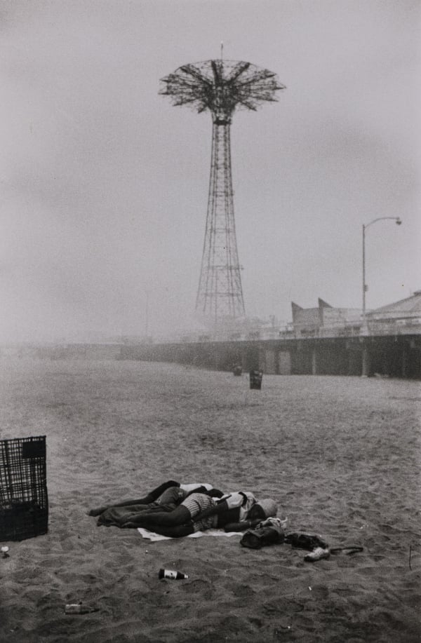 A photograph of the beach at Coney Island, showing people lying on the sand and an amusement park in the distance