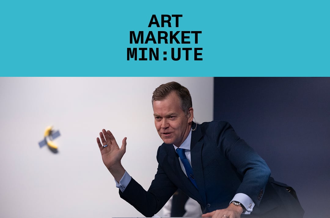 A man in a suit gestures energetically while speaking at an auction podiu, with a banana taped to the wall in the background. Text above reads "Art Market Minute."