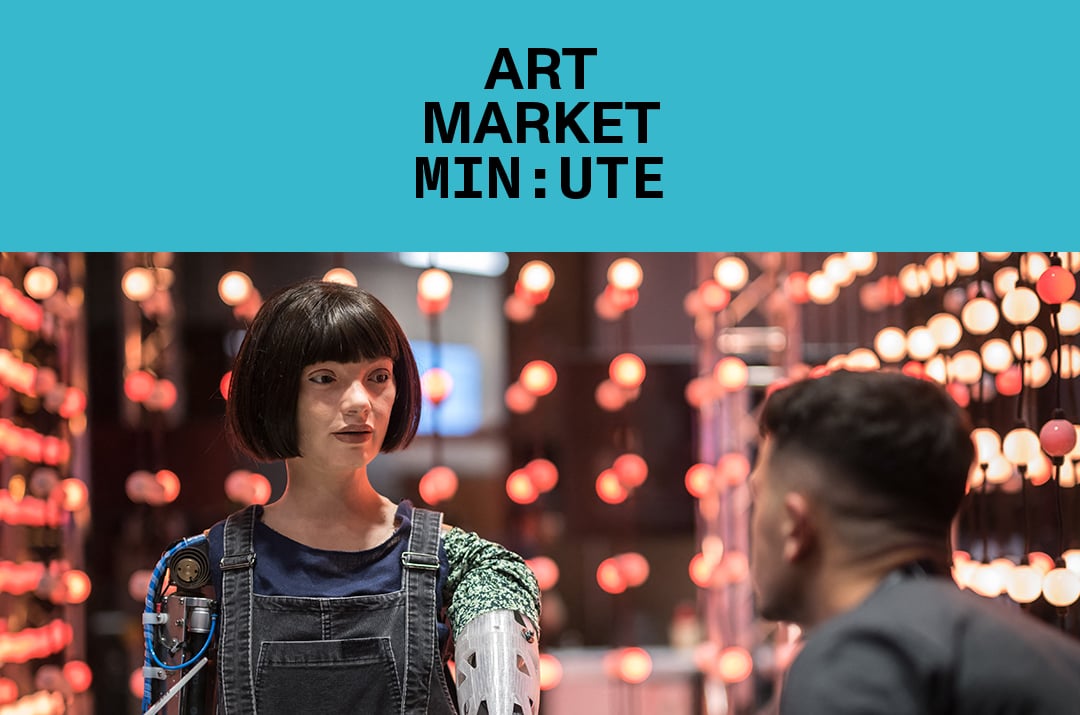 A lifelike robot with short dark hair interacts with a person in a setting illuminated by hanging pink and white lights, under a banner that reads "Art Market Minute."