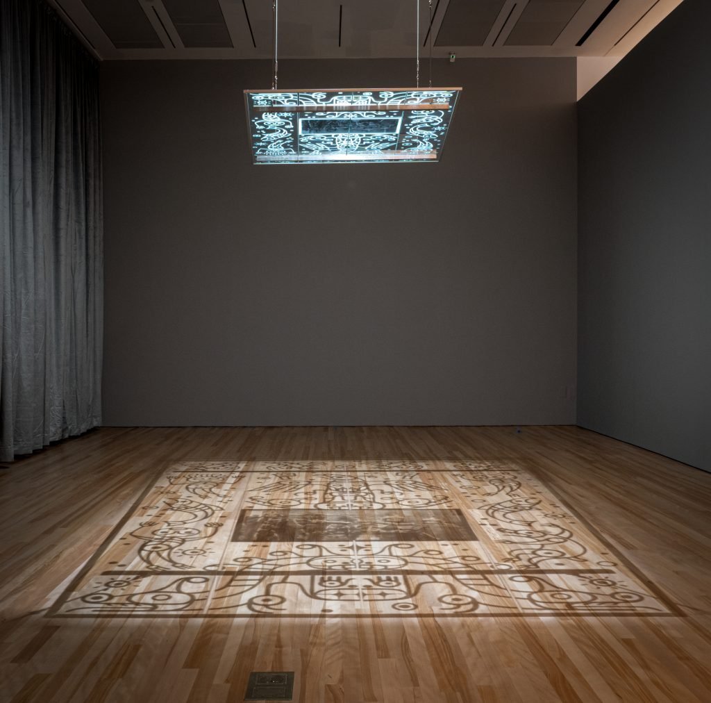 A dimly lit gallery installation featuring a suspended, illuminated panel projecting intricate designs onto the wooden floor below. The projections form a rectangular pattern with decorative, symmetrical motifs. The backdrop is a plain dark wall with curtains to one side, emphasizing the light and shadow effect of the artwork.