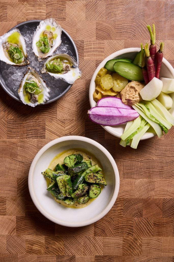 four dressed oysters sit atop an iced metal plate and two other plates contain colorful vegetable appetizers 