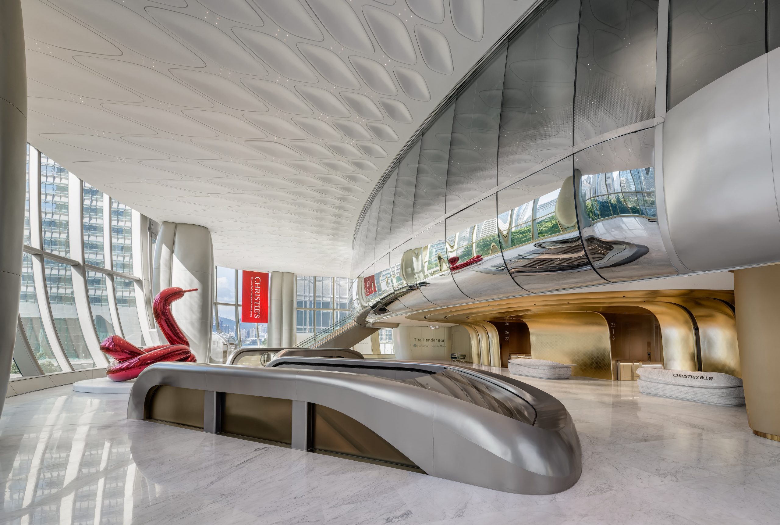 A futuristic looking of a building's lobby, with curved walls in silver on the right, a curved cover of an escalator on the front in silver. The floor to ceiling glass windows on the left. A large sculpture that appears to be a balloon swan is on the left. A banner that says Christie's in white on a red banner is hung vertically at the end of the lobby.