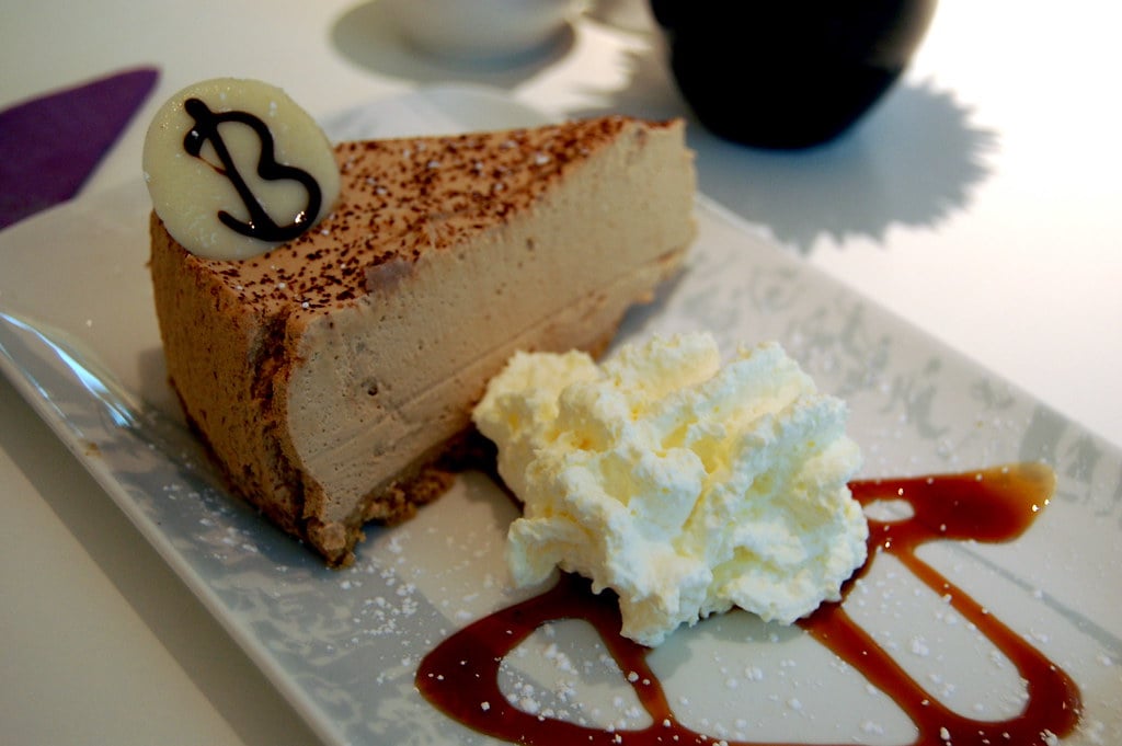 A piece of brown cake sits on a plate next to whipped cream.