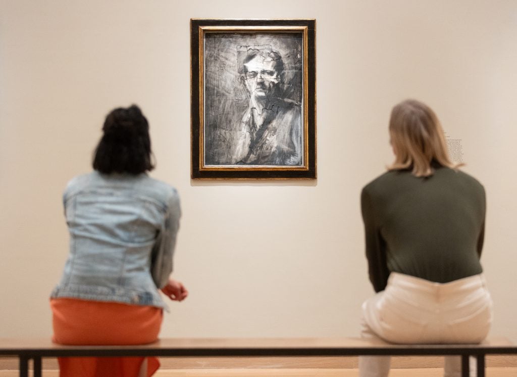 two people sit on a bench in an art gallery looking at a charcoal drawing framed on the wall