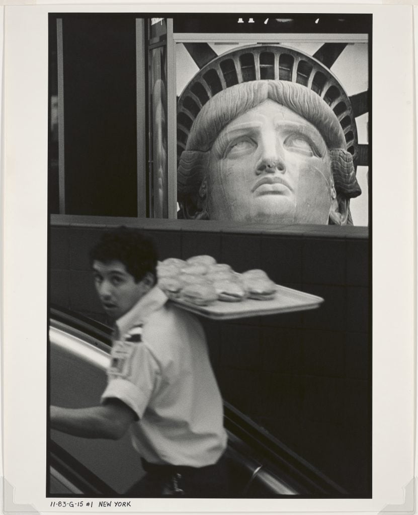 a black and white photo of a man carrying a tray of donuts with the head of the statue of liberty in the background