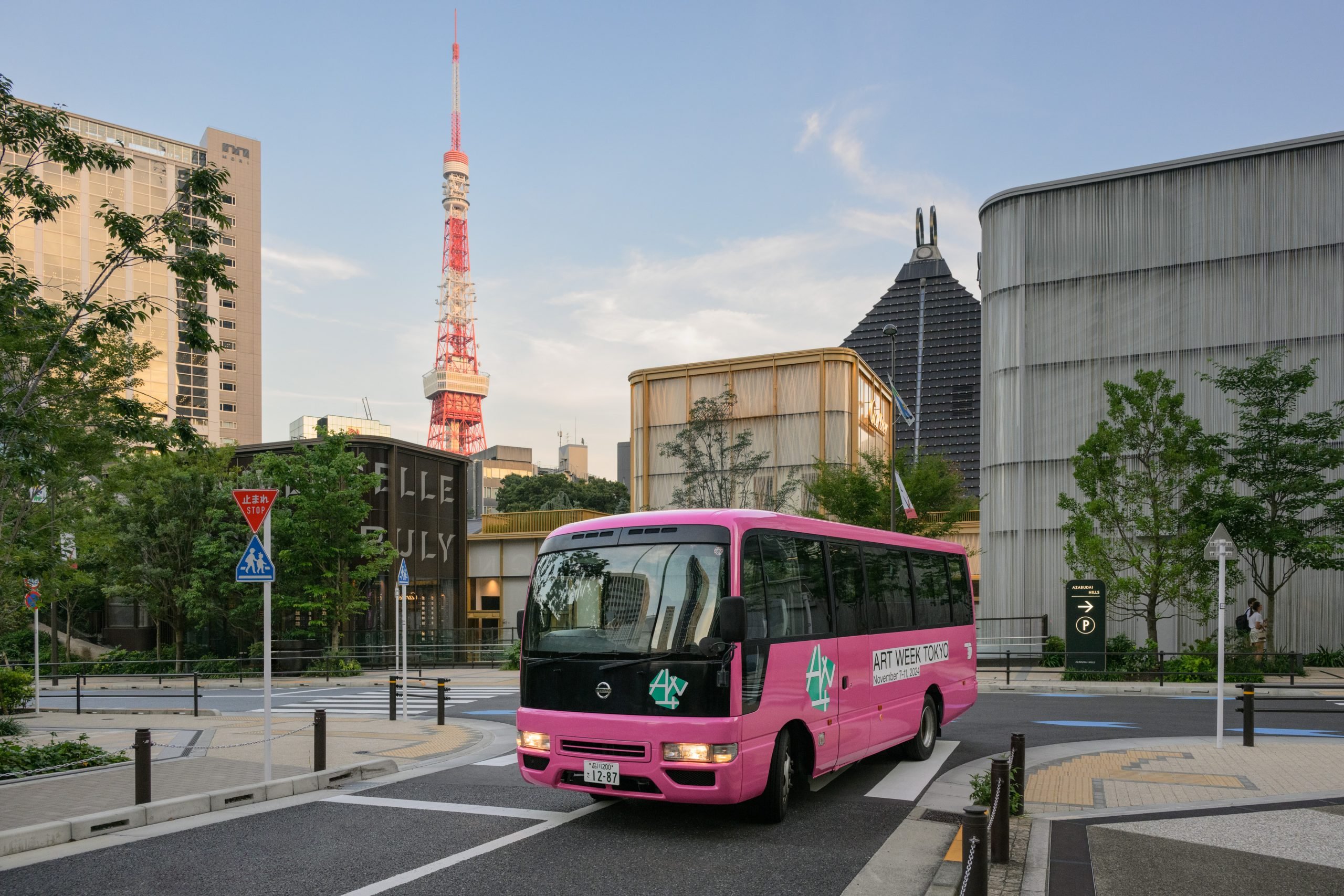 A pink mini-bus parked in the street surrounded by a grey building, some green bushes, and the Tokyo Tower at the back, under a blue sky