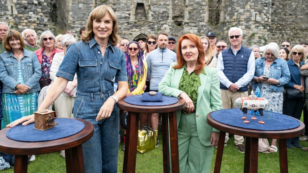 A crowd stand in front of a castle door with three raised wooden tables showcasing antique items.