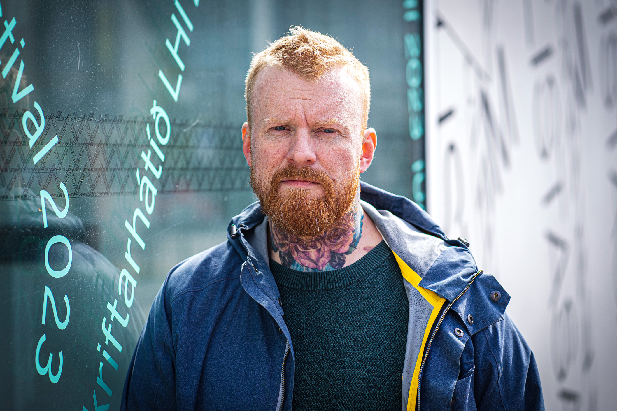 a man with a beard, a neck tattoo and in a blue jacket stands outside against a wall with writing