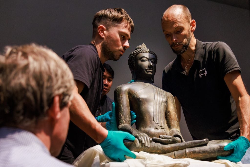 Two workers in black tee shirt and turquoise gloves carefully holding a buddha statue