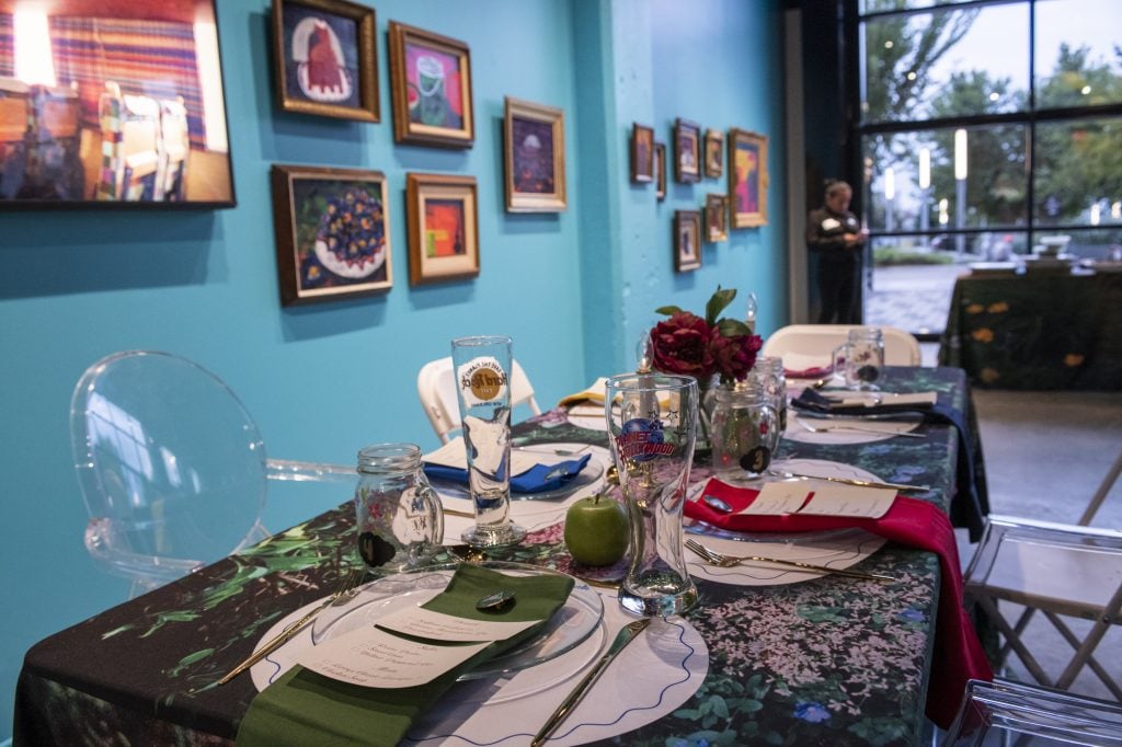 Azikiwe Mohammed designed the tablescapes for the opening reception for his show at Project for Empty Space Ironside. The tablecloth features a photograph by the artist, and there are many plates and glasses and flower arrangements on the table, with the artist's framed still life paintings hung on the robin's egg blue wall in gold frames.