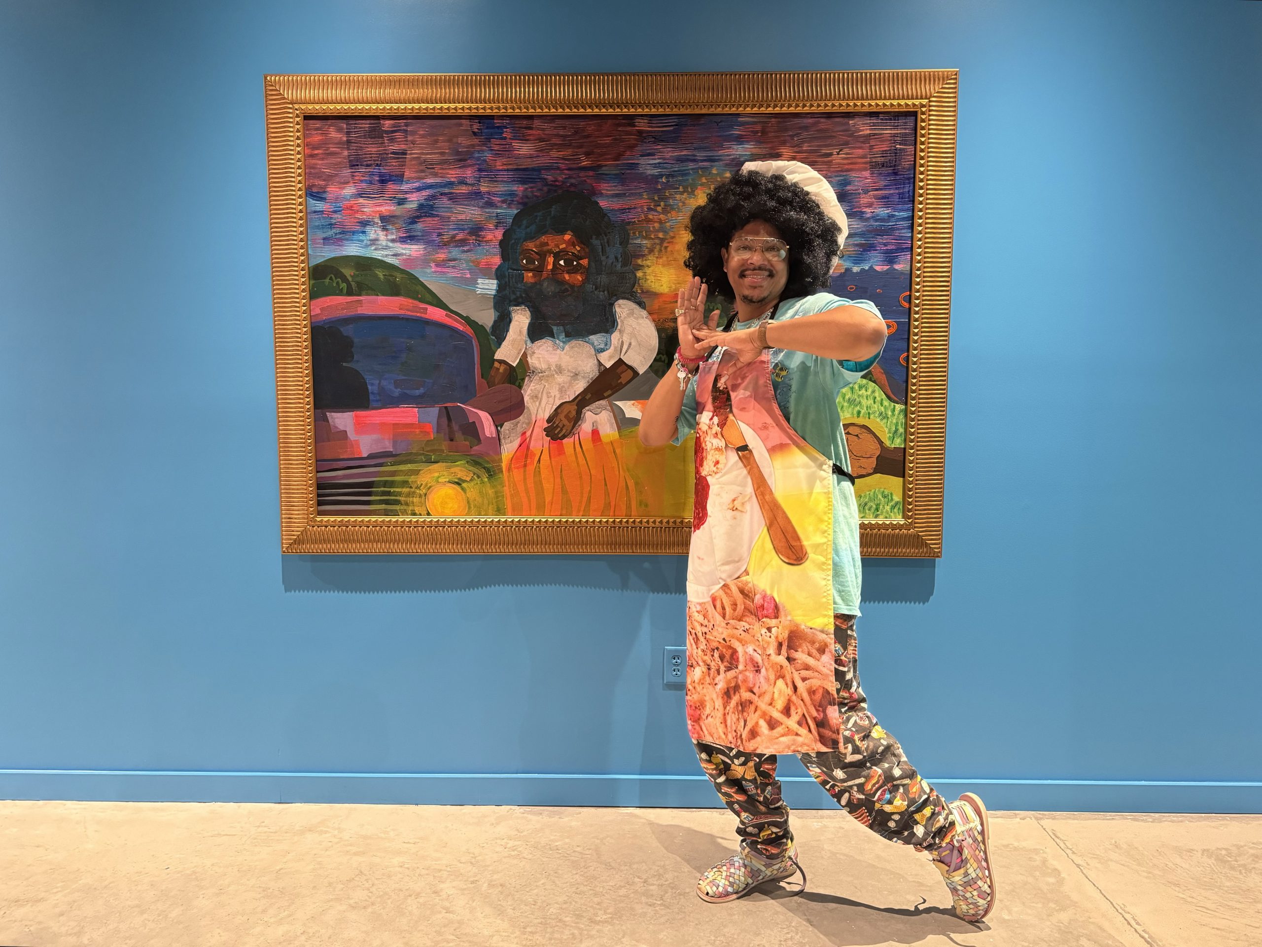 Azikiwe Mohammed, in costume as his "Leroy's Luncheon" character, at his show "Trains, Buses, and the Four C’s" at Project for Empty Space Ironside, Newark. He is a young Black man with a large afro dressed in a colorful outfit with an apron and chef's hat, in front of a large painting of a black woman in a hijab against a bright robin's egg blue wall.