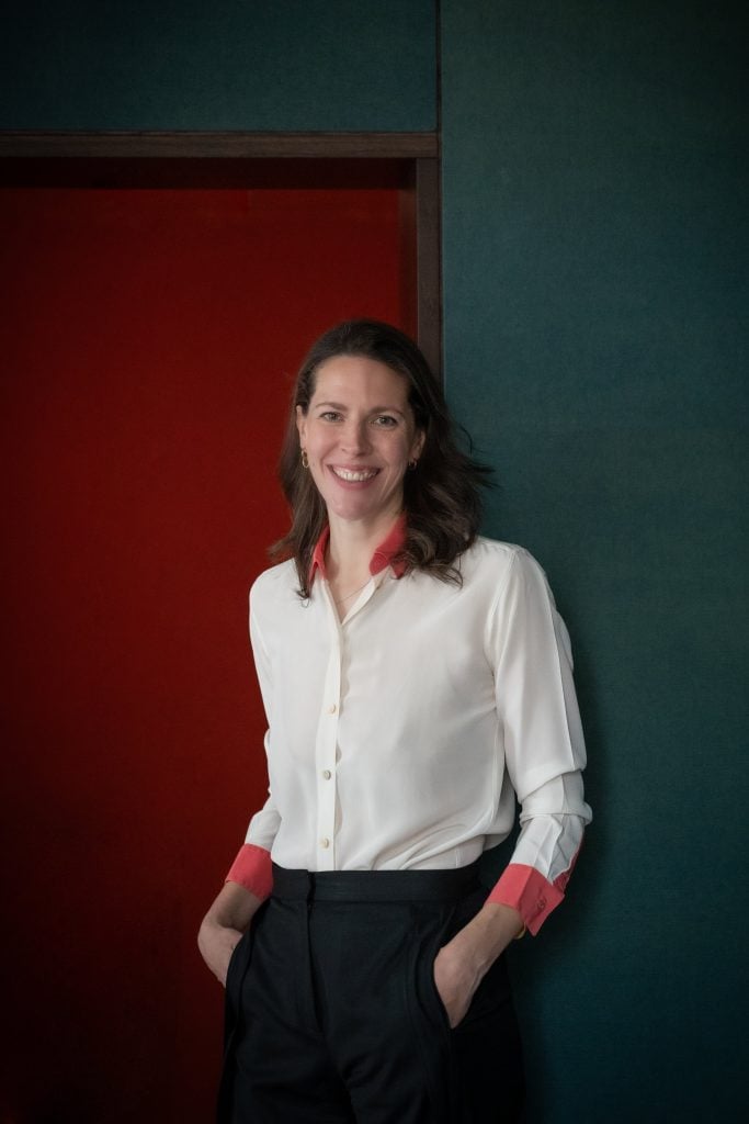 a woman with long dark hair stands in front of a minimalist painting wearing a white blouse 