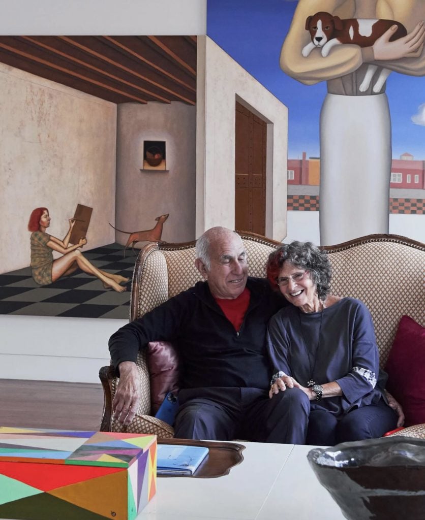 an older couple sitting on a sofa and smiling with large paintings behind them